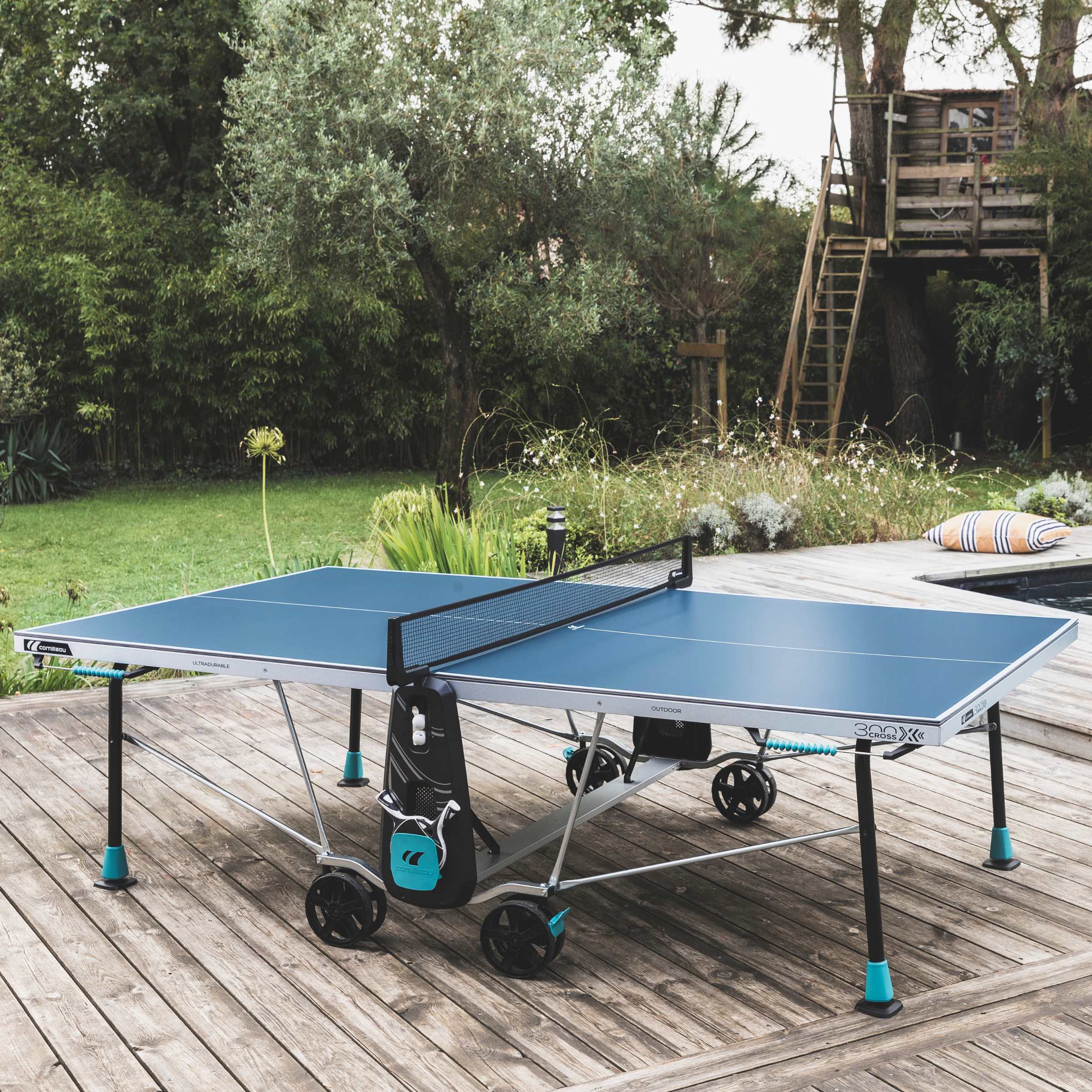 A blue portable table tennis table with wheels and a black net, set up on a wooden deck in a backyard with trees, grass, and a treehouse visible in the background.
