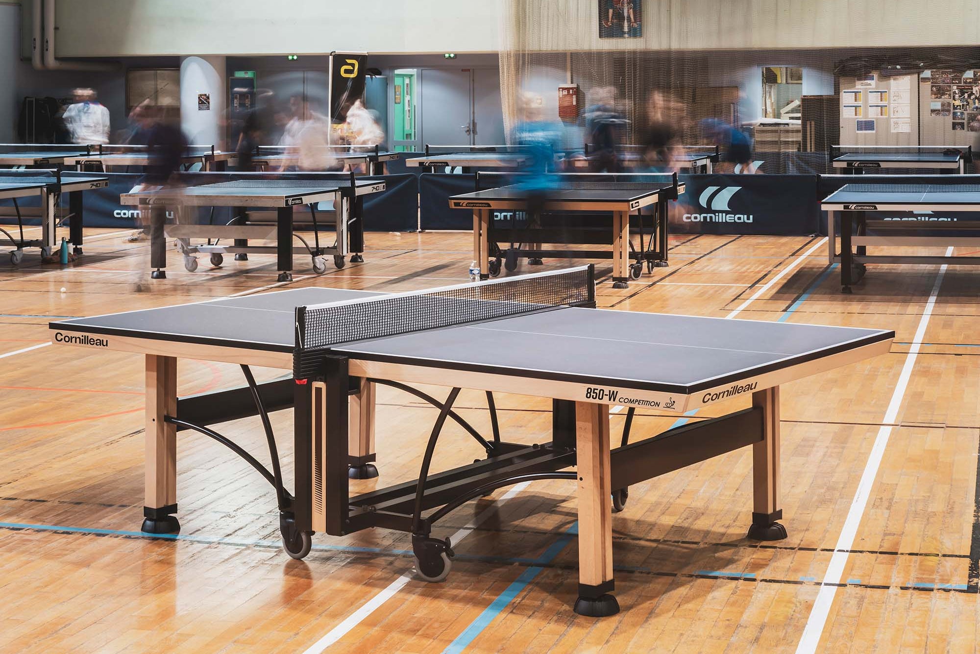 A Corbièu 250 table tennis table in the foreground of a large indoor sports hall with wooden floors, other tables, and blurred players in the background.